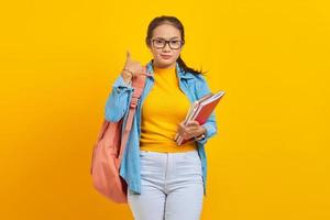 porträt einer lächelnden jungen asiatischen studentin in jeanskleidung mit rucksack, der notizbuch hält und eine telefongeste macht, wie sagt, ruf mich zurück, isoliert auf gelbem hintergrund foto