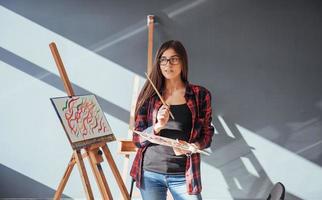 dunkelhaariger künstler hält einen pinsel in der hand und zeichnet ein bild auf leinwand. denkt, dass ziehen auf foto