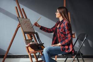 hübsche hübsche Künstlerin malt auf Leinwand auf der Staffelei. Studio schwarzer Hintergrund. lange haare, brünett. foto