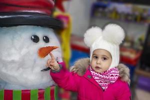 kleines Mädchen mit Schneemann. charmantes süßes mädchen steht in der nähe von schneemann. Winterferien für Kinder. foto