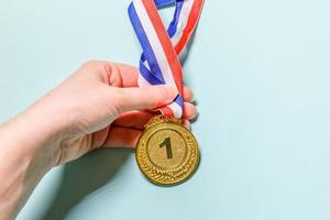 einfach flach gelegte Designhand, die Gewinner oder Champion-Goldtrophäenmedaille isoliert auf blauem, farbenfrohem Hintergrund hält. Sieg erster Platz des Wettbewerbs. gewinn- oder erfolgskonzept. Kopierbereich von oben. foto