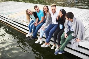 Porträt glücklicher junger Freunde, die auf einem Pier am See sitzen foto