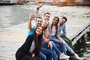 Gruppe schöner junger Leute, die Selfies machen, die auf dem Pier liegen, die besten Freunde von Mädchen und Jungen mit Vergnügen Konzept schafft emotionales Leben der Menschen.