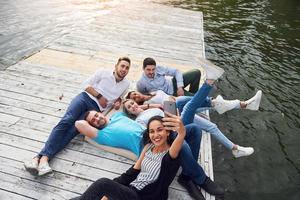 Gruppe schöner junger Leute, die Selfies machen, die auf dem Pier liegen, die besten Freunde von Mädchen und Jungen mit Vergnügen Konzept schafft emotionales Leben der Menschen. foto