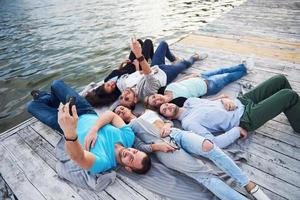 Gruppe schöner junger Leute, die Selfies machen, die auf dem Pier liegen, die besten Freunde von Mädchen und Jungen mit Vergnügen Konzept schafft emotionales Leben der Menschen. foto