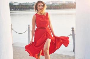 Fröhliche, schöne junge Frau, die im Sommer mit See im Hintergrund der Freiheit tanzt. Farbkontrast rot und weiß foto