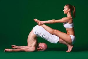 Schöne sportliche Frau und Mann in weißen Kleidern, die gemeinsam Yoga-Asanas im Innenbereich auf grünem Hintergrund machen foto