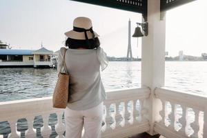 touristische frau, die spaß hat, während sie das stadtbild von bangkok neben dem fluss besichtigt, rückansicht einer asiatischen touristenfrau, die sich entspannt und genießt, während sie architektur auf ihrem handy fotografiert. foto