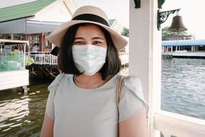 lächelnde touristische frau, die spaß hat, während sie das stadtbild von bangkok neben dem fluss besichtigt, porträt einer glücklich lächelnden touristischen frau, die während einer neuen normalen covid-19-situation eine gesichtsmaske trägt. reisekonzept foto