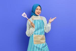 Fröhliche schöne asiatische muslimische Frau mit Schürze, Spachtel und ausgebreiteten Händen bei Hausarbeit isoliert über violettem Hintergrund. Housekeeping-Konzept foto
