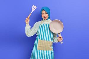 Fröhliche, schöne asiatische Frau, die eine Schürze trägt, die Pfannenwender und Bratpfanne hält, während sie Hausarbeiten isoliert auf violettem Hintergrund erledigt. Housekeeping-Konzept foto