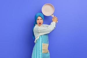 Wütende junge Asiatin in weißem Pullover mit Schürze und Hijab halten Bratpfanne, greifen Feinde an, während sie Hausarbeit isoliert über violettem Hintergrund machen. Housekeeping-Konzept foto