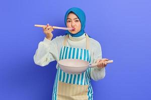 Fröhliche schöne asiatische Frau in weißem Pullover mit Schürze und Hijab, die Pfanne hält und Essen mit Löffel isoliert über violettem Hintergrund probiert. Housekeeping-Konzept foto