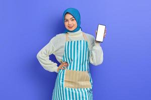 Porträt einer lächelnden jungen asiatischen muslimischen Frau mit Hijab und Schürze, die ein Handy mit leerem Bildschirm isoliert auf violettem Hintergrund hält. Menschen Hausfrau muslimischen Lifestyle-Konzept foto