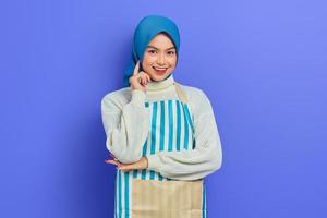 Porträt einer lächelnden jungen Hausfrau in Hijab und gestreifter Schürze, Finger auf die Wange legen, isoliert auf violettem Hintergrund. Menschen Hausfrau muslimischen Lifestyle-Konzept foto