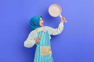 Porträt einer aufgeregten jungen asiatischen muslimischen Hausfrau, die eine Schürze trägt, die einen Pfannenwender hält und eine Bratpfanne isoliert auf violettem Hintergrund betrachtet. Housekeeping-Konzept foto