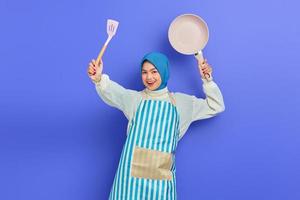 Fröhliche, schöne asiatische Frau in weißem Pullover, die mit Schürze und Hijab bedeckt ist, hält Pfannenwender und greift Feinde an, während sie Hausarbeit isoliert über violettem Hintergrund macht. Housekeeping-Konzept foto