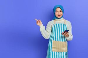 porträt einer lächelnden jungen asiatischen muslimischen frau, die hijab und schürze trägt, handy hält und mit den fingern isoliert über violettem hintergrund zur seite zeigt. Menschen Hausfrau muslimischen Lifestyle-Konzept foto
