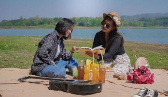 zwei glückliche junge asiatische frauen, die ein buch lesen, während sie am see urlaub machen foto