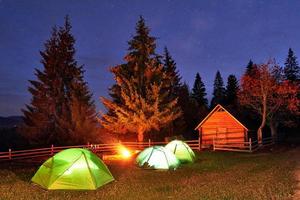 Nachtcamping. touristen ruhen sich an einem lagerfeuer in der nähe von beleuchtetem zelt und holzhaus unter einem erstaunlichen nachthimmel voller sterne und milchstraße aus. Astrofotografie foto