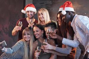 Gruppe schöne junge Leute, die Selfie in der Neujahrsparty machen, beste Freunde, Mädchen und Jungen, die zusammen Spaß haben und emotionale Lifestyle-Leute posieren. Hüte Weihnachtsmänner und Sektgläser in den Händen foto