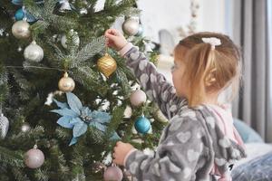 frohe weihnachten und schöne feiertage. Junges Mädchen, das beim Schmücken des Weihnachtsbaums hilft und einige Weihnachtskugeln in der Hand hält foto