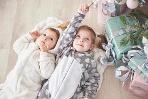 Zwei süße kleine Mädchen im Schlafanzug, die unter den Geschenken unter dem Weihnachtsbaum liegen