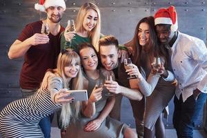 gruppieren sie schöne junge leute, die selfie auf der neujahrsparty machen, beste freunde mädchen und jungen, die zusammen spaß haben, lebensstilmenschenkonzept posieren. hüte weihnachtsmänner und champagnergläser in ihren händen foto