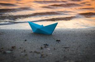 Das papierblaue Schiff wird bei Sonnenuntergang auf das sandige Ufer des Sees geworfen.