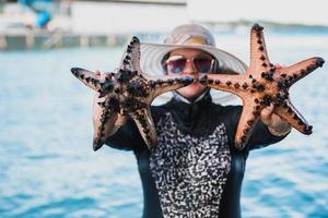 glückliche Frau mit Sonnenbrille, die Seesterne mit Meereshintergrund bei Karimun Jawa zeigt und hält foto