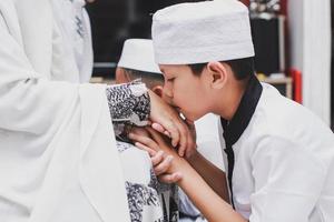 asiatischer muslimischer junge mit traditioneller kleidung, der zu hause bei der feier von eid mubarak die hand der mutter küsst foto