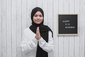 muslimische frau, die eine grußpose als symbol des verzeihens mit briefbrett macht, sagt glückliches eid mubarak an der wand foto