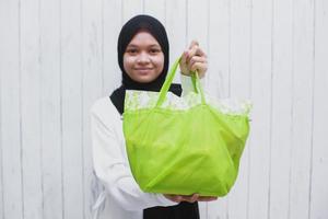 glückliche asiatische muslimische frau, die eid-geschenke in einer grünen tasche zeigt und gibt foto