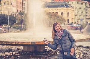 junge Touristin mit grauer Jacke posiert und hält therapeutisches Mineralwasser in der Tasse in der Nähe der heißen Quelle des Brunnens foto