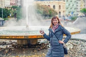 junge Touristin mit grauer Jacke lächelt und hält therapeutisches Mineralwasser foto