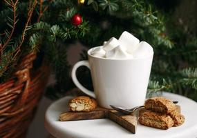 Kaffee in einer weißen Tasse mit Marshmallows. morgendlicher festlicher kaffee mit traditionellen italienischen cantuccini-mandelkeksen. eine tasse kaffee auf einem hintergrund grüner tannenzweige auf einem weißen stand. foto