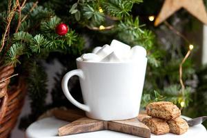 Kaffee in einer weißen Tasse mit Marshmallows. morgendlicher festlicher kaffee mit traditionellen italienischen cantuccini-mandelkeksen. eine tasse kaffee auf einem hintergrund grüner tannenzweige auf einem weißen stand. foto