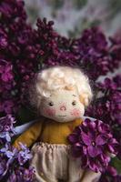 kleiner goldhaariger Engel in den blauen, rosa, purpurroten, violetten lila Blumen. handgefertigtes Spielzeug in violett-lila Farben. Grußkarte.