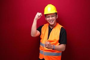 Fröhlicher, aufgeregter und lächelnder junger asiatischer Arbeiter, der seinen Arm hochhebt, um Erfolg oder Leistung zu feiern. foto