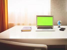 Laptop-Computer mit grünem Bildschirm, der auf einem Heimarbeitstisch neben Notebook, Frühlingsblumen und Weltkugelständer sitzt. konzept minimalistischer digitaler studentenarbeitsplatz foto
