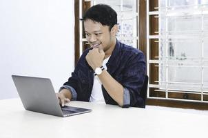 junger asiatischer mann, der sich glücklich fühlt und lächelt, wenn der arbeitslaptop auf dem tisch liegt. indonesischer Mann mit blauem Hemd. foto