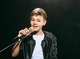 Porträt eines kaukasischen Teenagers in weißem T-Shirt, Blue Jeans und Lederjacke mit Mikrofon, das auf dunklem Hintergrund singt. hobby- und ruhmkonzept foto