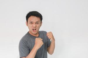 Wütendes und hasses Gesicht des jungen asiatischen Mannes im grauen T-Shirt mit wütender Handbewegung foto