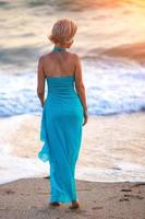 Ein junges schlankes Mädchen steht bei Sonnenuntergang in einem blauen langen Kleid am Strand, eine schöne Figur foto