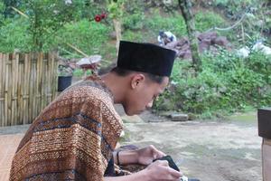 14. märz 2022 in cianjur regency, west java, indonesien. Foto von der Seite eines jungen Mannes, der indonesische Kleidung trägt und ein Smartphone hält. er spielt ein Spiel.