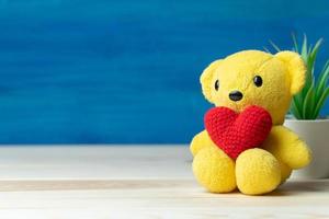 Handgemachtes Garn Rotes Herz auf gelbem Teddybär vor weißem Topf und grünen Zierpflanzen auf Holztisch und blauem Hintergrund. Konzept des Valentinstags.