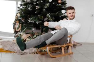 Fröhlicher Mann, der zu Hause auf dem Schlitten in der Nähe des Weihnachtsbaums sitzt. Mann auf einem Schlitten. Weihnachten gute Laune. familien- und urlaubskonzept. foto