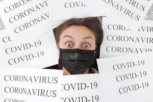 schockierte junge schöne frau in medizinischer schwarzer maske liegt in papieren mit der aufschrift coronavirus und covid-19. Quarantäne zu Hause. selektiver Fokus foto