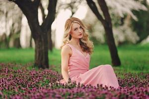 schöne blonde junge Frau in bunten Blumen. Mädchen mit Make-up und Frisur im rosafarbenen Kleid im blühenden Frühlingspark. Frauentag foto