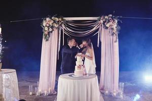 wunderschöne Braut und stilvoller Bräutigam probieren ihre stilvolle Hochzeitstorte. glückliches frisch verheiratetes paar, das stück kuchen isst, lustiger emotionaler moment foto